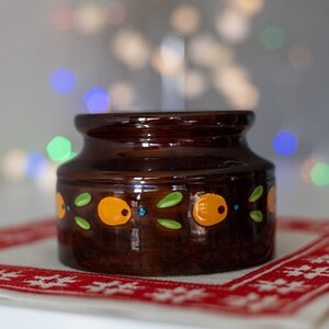 Può includere: Un piccolo vaso di ceramica marrone con finitura lucida. Il vaso è decorato con disegni dipinti di arancione, verde e blu. È appoggiato su una superficie bianca e rossa a motivi. Lo sfondo è sfocato con luci bokeh colorate.
