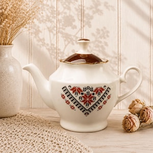May include: A cream-colored ceramic teapot featuring a dark brown lid with a gold-toned rim. The body of the teapot is decorated with a geometric pattern in red, black, and gray, consisting of star-like shapes and linear borders.