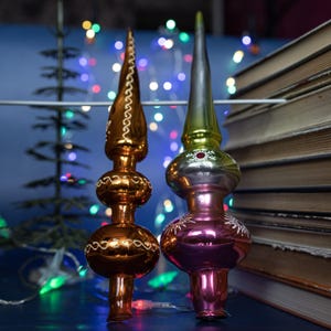 May include: Two vintage Christmas tree toppers. One is gold with white accents, the other is silver, green, and pink with snowflake designs. The toppers are glass and have a classic, ornate design. A stack of books and string lights are in the background.