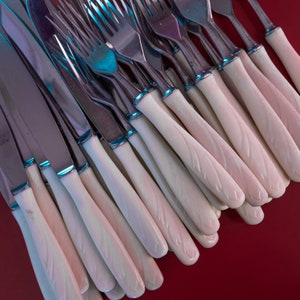 May include: Close-up of a collection of silverware, including knives and forks. The silverware features stainless steel blades and tines, with off-white handles. The handles have a textured, leaf-like design. The silverware is arranged on a red surface.