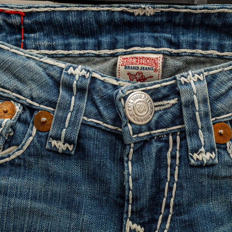 Puede incluir: Primer plano de unos vaqueros de mezclilla azules con costuras blancas. Los vaqueros tienen cierre de bot&oacute;n, remaches de cobre y una etiqueta de la marca True Religion. Los detalles de las costuras son prominentes.