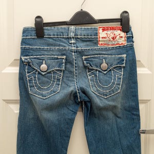 Puede incluir: Vaqueros de mezclilla azul con costuras blancas y detalles de botones plateados. Los bolsillos traseros presentan un dise&ntilde;o de herradura. Es visible un parche rojo y blanco de la marca True Religion.
