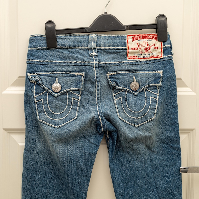 Puede incluir: Vaqueros de mezclilla azul con costuras blancas y detalles de botones plateados. Los bolsillos traseros presentan un dise&ntilde;o de herradura. Es visible un parche rojo y blanco de la marca True Religion.