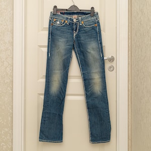 Puede incluir: Vaqueros azules de mezclilla con un lavado descolorido y costuras blancas. Los vaqueros tienen un dise&ntilde;o cl&aacute;sico de cinco bolsillos, cierre de bot&oacute;n y cremallera, y cuelgan de una percha negra. Los vaqueros tienen una ligera abertura de pierna acampanada.