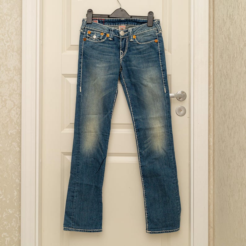 Puede incluir: Vaqueros azules de mezclilla con un lavado descolorido y costuras blancas. Los vaqueros tienen un dise&ntilde;o cl&aacute;sico de cinco bolsillos, cierre de bot&oacute;n y cremallera, y cuelgan de una percha negra. Los vaqueros tienen una ligera abertura de pierna acampanada.