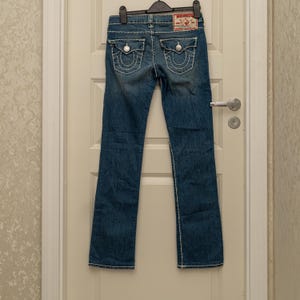 Puede incluir: Vaqueros de mezclilla azul con costuras blancas y detalles en los bolsillos traseros. Cuenta con un parche de marca rojo y blanco en la parte trasera. Los vaqueros est&aacute;n colgados en una percha negra.