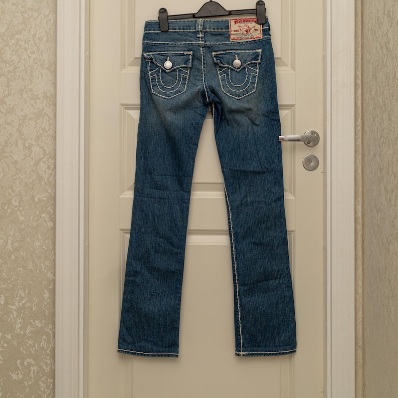 Puede incluir: Vaqueros de mezclilla azul con costuras blancas y detalles en los bolsillos traseros. Cuenta con un parche de marca rojo y blanco en la parte trasera. Los vaqueros est&aacute;n colgados en una percha negra.