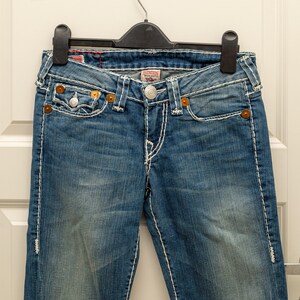 Puede incluir: Vaqueros azules de mezclilla con costuras blancas, cierre de bot&oacute;n y bolsillos delanteros y traseros. Los vaqueros est&aacute;n colgados de una percha negra. Los vaqueros tienen un lavado descolorido y son de estilo cl&aacute;sico.