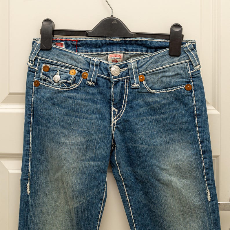 Puede incluir: Vaqueros azules de mezclilla con costuras blancas, cierre de bot&oacute;n y bolsillos delanteros y traseros. Los vaqueros est&aacute;n colgados de una percha negra. Los vaqueros tienen un lavado descolorido y son de estilo cl&aacute;sico.