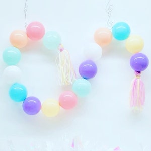 May include: A pastel colored garland made of balloons with tassels. The garland is strung together with wire and has a variety of colors including pink, blue, purple, yellow, and white.
