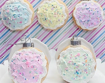 May include: Five round cookie-shaped ornaments with textured tan edges and pastel-colored frosting tops. Each ornament features white piped icing borders and colorful sprinkles in shades of blue, yellow, lavender, pink, and mint green.