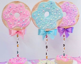 Op de afbeelding: Drie donutvormige tafeldecoraties met roze, blauwe en gele glazuur. Elke donut is versierd met witte glazuur en kleurrijke sprinkels. De donuts zijn bevestigd aan witte rietjes met zwarte en witte donutpatronen. Elk rietje is versierd met een strik in roze, blauw en paars.