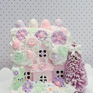 Puede incluir: Una casa rosa pastel con temática de Pascua con glaseado blanco, flores decorativas y detalles de huevo. También se incluye un pequeño árbol morado y un conejito verde. La casa tiene ventanas negras y un acabado brillante.