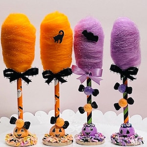 May include: Three Halloween-themed cotton candy trees with black ribbon bows. The trees are decorated with candy, sprinkles, and Halloween-themed characters. The cotton candy is orange, purple, and pink.