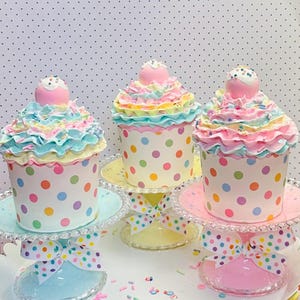 May include: Three decorative cupcakes with pastel frosting and sprinkles sit on glass stands with ribbon bows. The cupcake wrappers are white with colorful polka dots. Each cupcake is topped with a pink ball.