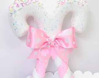 May include: White candy cane ornament with pink and white stripes, topped with white glitter and pastel sprinkles. A pink polka dot bow is tied in the center.