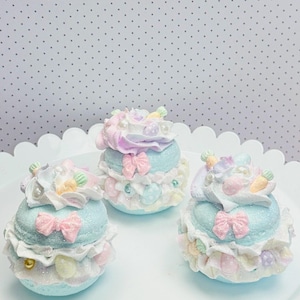 May include: Three pastel-colored decorative cupcake-shaped objects. Each has a light blue base, white frills, and a swirl of pastel pink and purple frosting. They are adorned with pink bows, faux pearls, and small carrot and egg decorations. The objects are on a white scalloped tray.