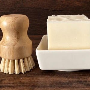 May include: A light-colored wooden dish brush with stiff bristles positioned next to a square white ceramic soap dish containing a solid, rectangular block of off-white soap. The brush features a rounded handle and a flat base for upright storage.