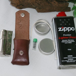 Puede incluir: Un encendedor plateado con una funda de cuero, un pequeño recipiente de fluido para encendedor, una mecha y un pequeño recipiente de piedras de pedernal. La botella de fluido para encendedor es negra con el texto "Zippo Premium Lighter Fluid" y "Cleaner Burning Improved Ignition Low Odor" en blanco. La botella también tiene una tapa roja.