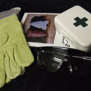 May include: A green work glove, a small white first aid kit with a green cross, safety glasses, and a small white card with a picture of a stone arrowhead.