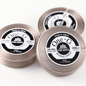 Könnte beinhalten: Drei runde Spulen mit hellbeigem One-G Perlenfaden. Jede Spule hat ein weißes Etikett mit schwarzem Text, der One-G, Toho Beads, 330 dtex, 100% Nylon und 250 yd (229 m) lautet. Der Faden ist fest um die runden Kunststoffspulen gewickelt.
