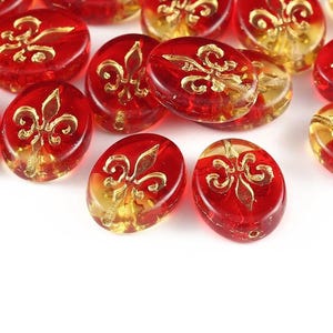 May include: A collection of oval-shaped glass beads featuring a translucent red and amber gradient finish. Each bead is decorated with a raised gold-coloured fleur-de-lis emblem centred on the front surface, creating a repeating pattern across the group.