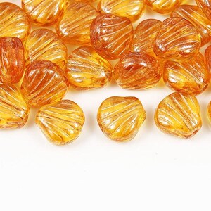 May include: A collection of translucent amber-colored glass beads shaped like small seashells. Each bead features a series of raised, radiating ridges on its surface, creating a textured, fan-like appearance with a glossy, reflective finish.