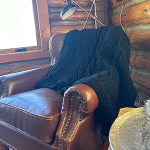 May include: A brown leather armchair with a black cable knit throw draped over it. The chair has a high back and a tufted seat. The chair is in a room with wood panelling on the walls.