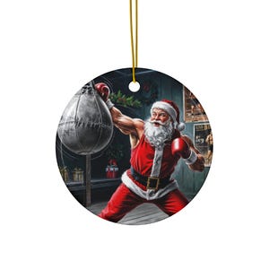 May include: Round Christmas ornament featuring Santa Claus in a boxing pose, wearing red boxing gloves and a red outfit with white trim. Santa is punching a gray punching bag. The background includes boxing posters and a wreath.