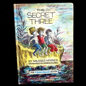 May include: A hardcover book titled The Secret Three by Mildred Myrick with drawings by Arnold Lobel. The cover features a colorful illustration of three children sitting on a cliff edge above water, with the text An I Can Read Book and Harper & Row Publishers.