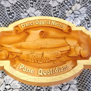 May include: A wooden bread board with a carved design of a loaf of bread, grapes, and a pitcher. The board has the text "Dacci Oggi il Nostro Pane Quotidiano" carved into it.