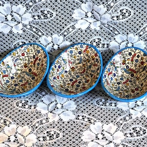 May include: Three oval-shaped ceramic bowls with a bright blue rim and a detailed interior pattern featuring small red, yellow, and blue floral motifs on a white background with thin, dark, winding vine-like lines connecting the flowers.