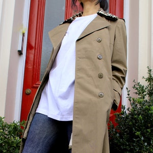 May include: A long, beige trench coat with a leopard print collar. The coat is worn over a white t-shirt and blue jeans.