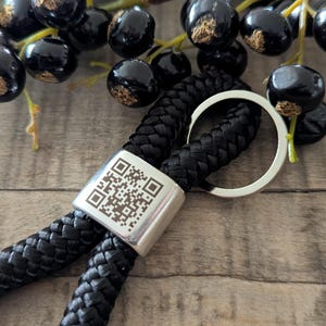 Könnte beinhalten: Ein schwarzes geflochtenes Schlüsselband mit einem silberfarbenen Metallverschluss mit einem QR-Code. Ein silberner Ring ist am Schlüsselband befestigt. Dunkle Beeren sind im Hintergrund.