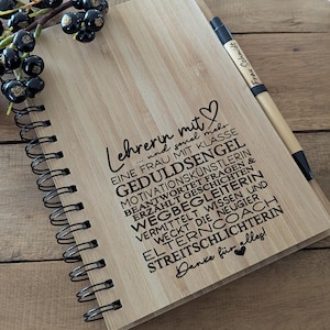 Könnte beinhalten: Ein holzfarbenes Notizbuch mit Spiralbindung und deutschem Text, darunter "Lehrerin mit...". Ein Stift mit der Aufschrift "Frau Schmidt" liegt daneben. Dunkle Beeren befinden sich in der oberen linken Ecke.