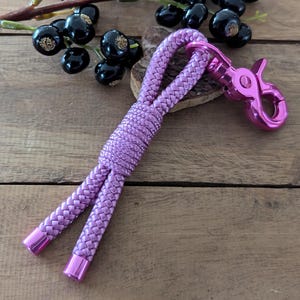 Könnte beinhalten: Ein pinkfarbener geflochtener Schlüsselband mit einem rosafarbenen Metallverschluss. Der Schlüsselband liegt auf einer Holzoberfläche.