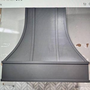 May include: A matte dark grey range hood with a curved, flared silhouette and vertical trim detailing. The structure features a wide, horizontal base band and a smooth, solid finish, designed for installation above a kitchen cooking area.