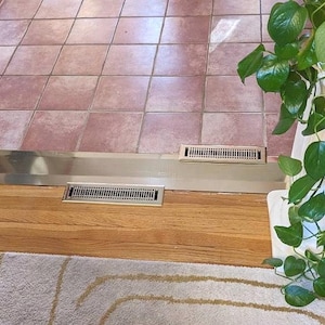 May include: Two rectangular, metallic air vents are set into a wooden floor, positioned below a metal strip. The floor transitions to a tiled surface. A green leafy plant is visible in the upper right corner, adding a touch of nature to the scene.