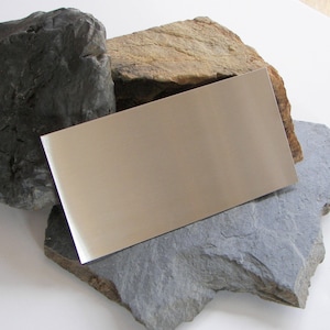 May include: A rectangular piece of polished metal with a reflective surface, resting on top of three different colored rocks.