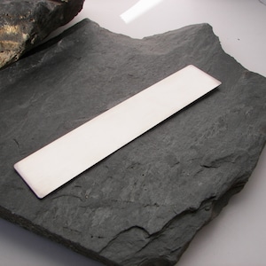 May include: A long, rectangular, silver bookmark on a dark gray, textured surface.