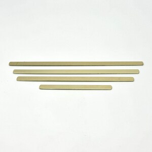 May include: Four flat, rectangular, metallic gold bars of varying lengths are arranged horizontally against a white background. The bars appear to be made of a brushed metal material, with a slightly reflective surface.