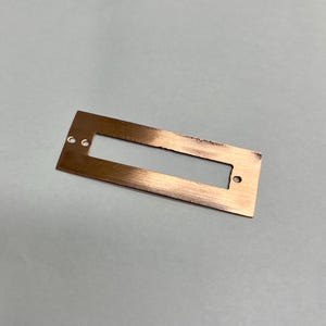 May include: A rectangular copper-colored metal plate with a long, narrow cutout in the center. The plate has two small holes on one end and one on the other. The metal has a brushed texture.