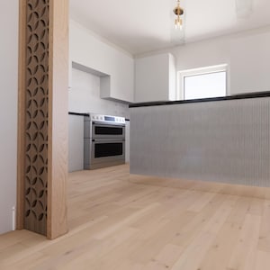 May include: A modern kitchen interior with light wood flooring and white walls. A large island with vertical paneling and a dark countertop is in the center. A decorative wooden pillar with a geometric cutout design is on the left.