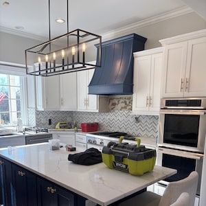 May include: A modern kitchen with white cabinets, a marble countertop island, and a navy blue range hood. A black rectangular light fixture hangs above the island. A yellow and black Ryobi tool box sits on the island.