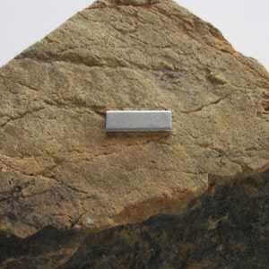 May include: A close-up of a small, rectangular, silver-colored metal bar resting on a rough, brown, textured rock surface.