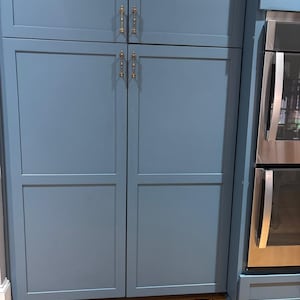 May include: Blue kitchen cabinets with gold-colored handles. The cabinets have a recessed panel design and are next to a stainless steel oven. The cabinets are above a wooden floor.