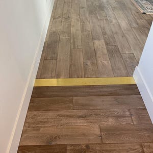 May include: Wooden flooring with a brass transition strip. The floorboards are a mix of light and dark brown tones, creating a natural wood grain pattern. The brass strip adds a touch of elegance and separates the two flooring sections.