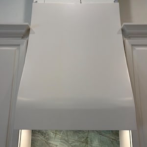 May include: A matte white kitchen range hood with a smooth, tapered design positioned between two white cabinets. The hood features a clean, minimalist front panel and is illuminated from underneath, casting light onto a green marble backsplash.
