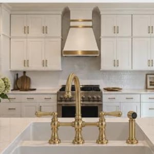 May include: A brass bridge-style kitchen tap with two lever handles and a side spray, mounted on a white farmhouse sink. The tap features a high-arc spout and a metallic finish, set against a kitchen with white cabinets and a range hood.