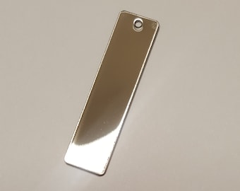 Puede incluir: Una etiqueta rectangular de metal plateado con un agujero en la parte superior. La etiqueta está en blanco y lista para ser grabada.